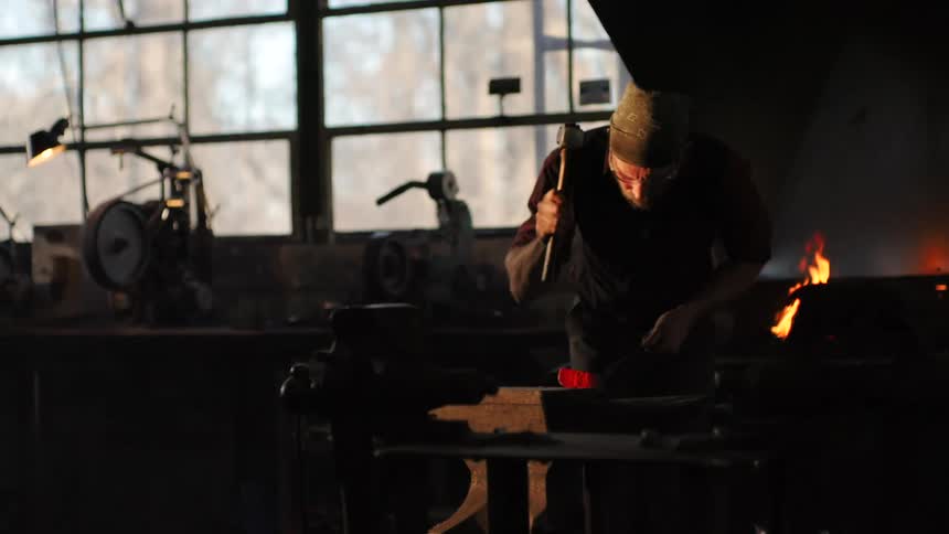 A blacksmith working in a dimly lit forge striking a heated piece of metal on an anvil while a fire burns in the background creating an atmospheric and industrial setting
