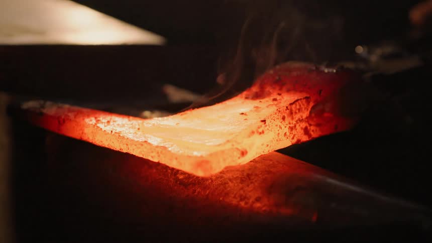 A blacksmith strikes hot metal on an anvil with a hammer holding it steady with tongs in a dimly lit workshop