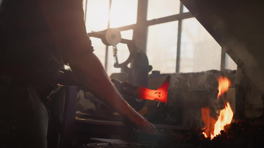 Blacksmith adds powder onto a glowing metal object while standing near the forge in his workshop on a sunny day
