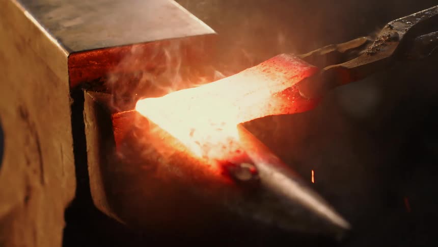 A closeup shows a hammer striking redhot iron held by tongs over an anvil