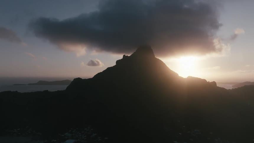 An aerial view at dusk flies over the ocean towards a silhouetted mountain peak partially obscured by a dramatic dark cloud formation light shining behind