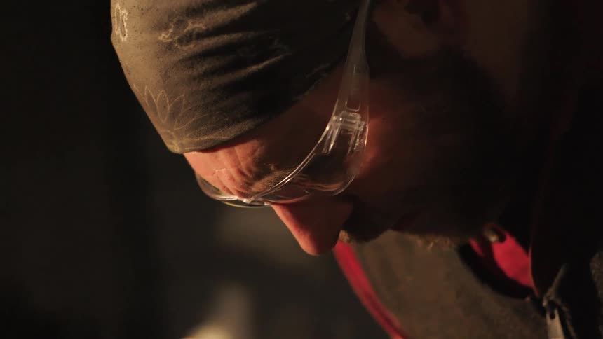 A closeup of a bearded man wearing a bandana and safety glasses as he hammers metal in a blacksmith workshop