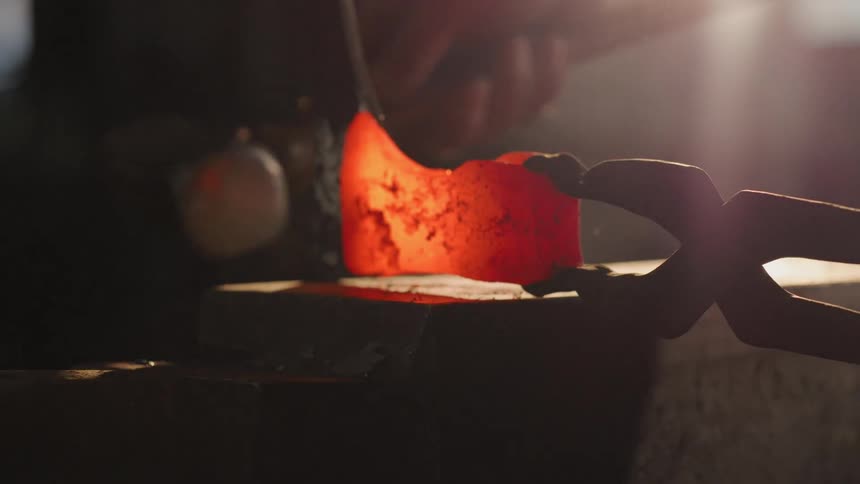 A glowing redhot piece of metal is gripped by tongs and struck with a hammer over an anvil set against a shadowy backdrop
