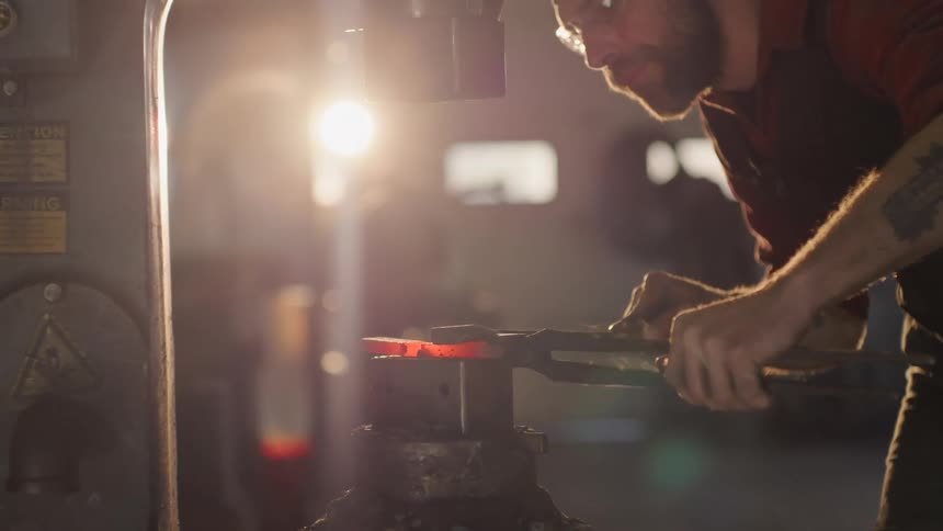 Man manipulates a heated metal part under the pressure of a press in a workshop