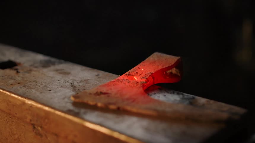 A blacksmith uses a hammer to strike a bolt held with visegrip pliers against a redhot blade causing a shower of bright metal sparks to fly off