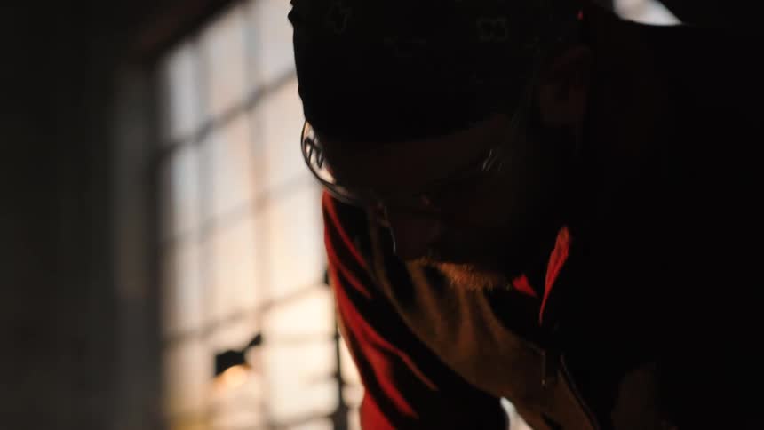 A bearded blacksmith wearing safety glasses and a bandana is intensely focused as he works with a hammer in a dimly lit workshop