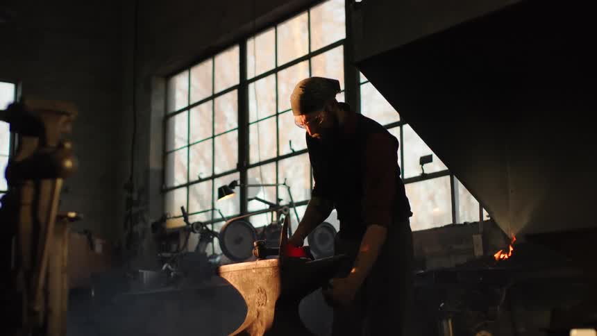 Male blacksmith manipulates a glowing metal object on an anvil during the day
