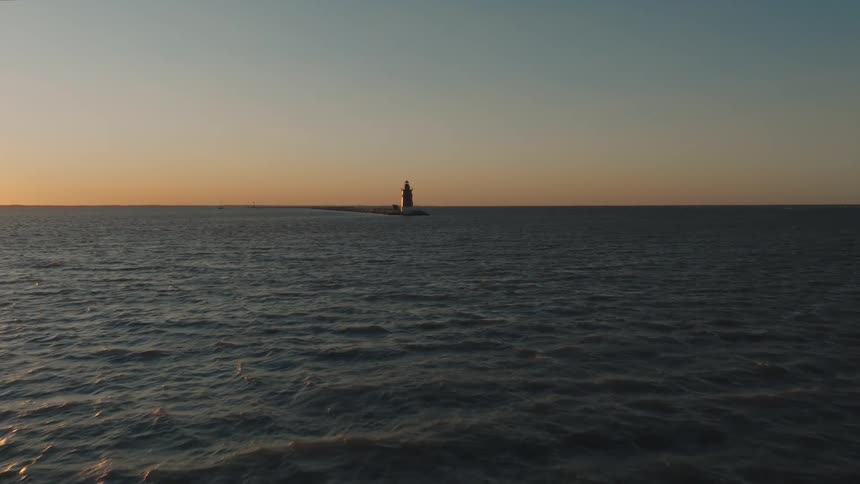An aerial view moves over the ocean towards a distant lighthouse standing on a rocky outcrop at sunset the water calm below