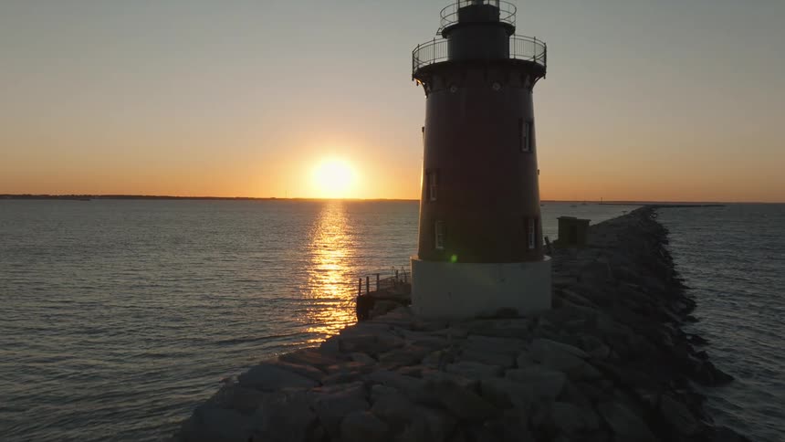 The sun is setting behind a lighthouse that is at the end of a long section of rocks