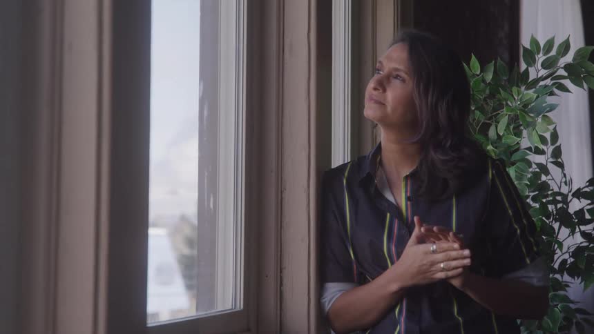 A woman stands by a window in a room rubbing her hands thoughtfully