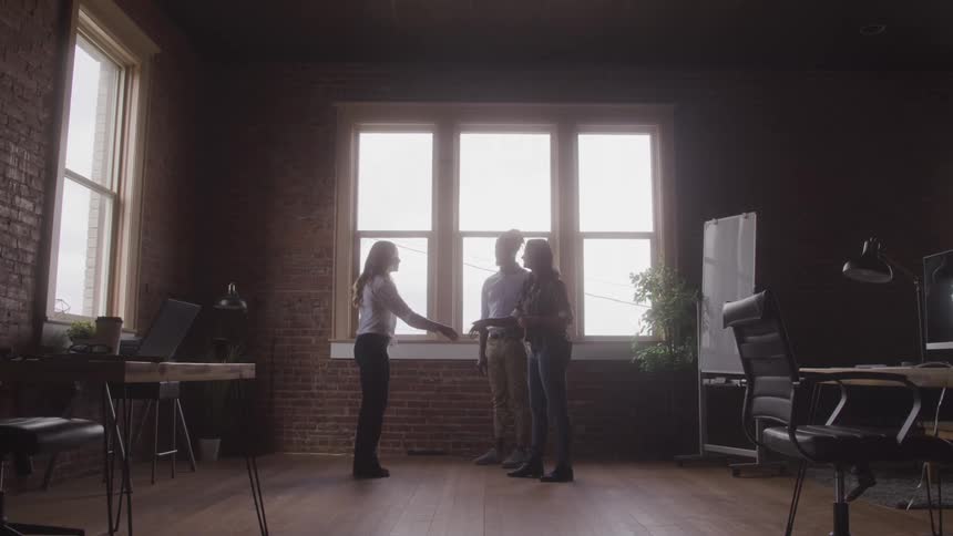 A woman shakes hands with two others in a modern workspace with large windows brick walls and wood floor