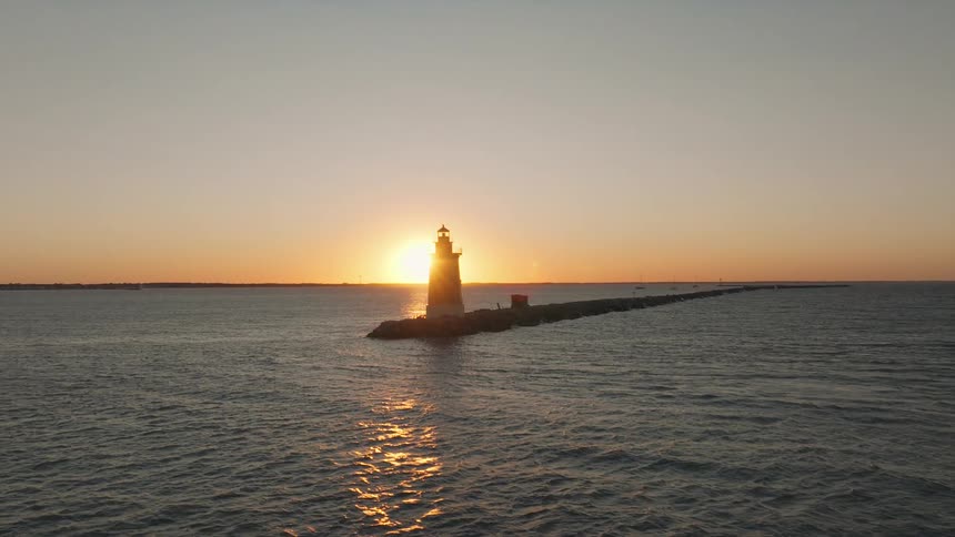A lighthouse stands silhouetted against a sunset sky with the suns rays bursting behind it and reflecting on the surrounding water