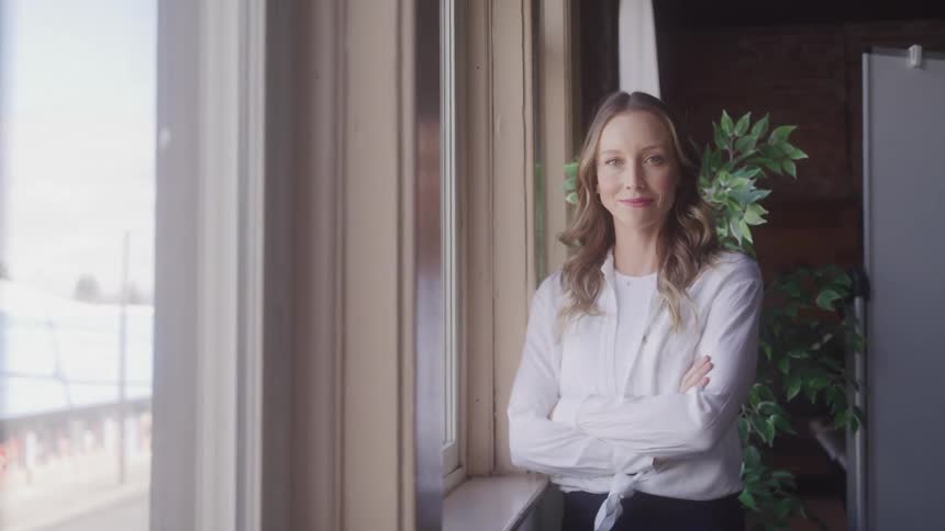 Girl with her arms folded looking out a window then turns to the camera convinced and with a smile