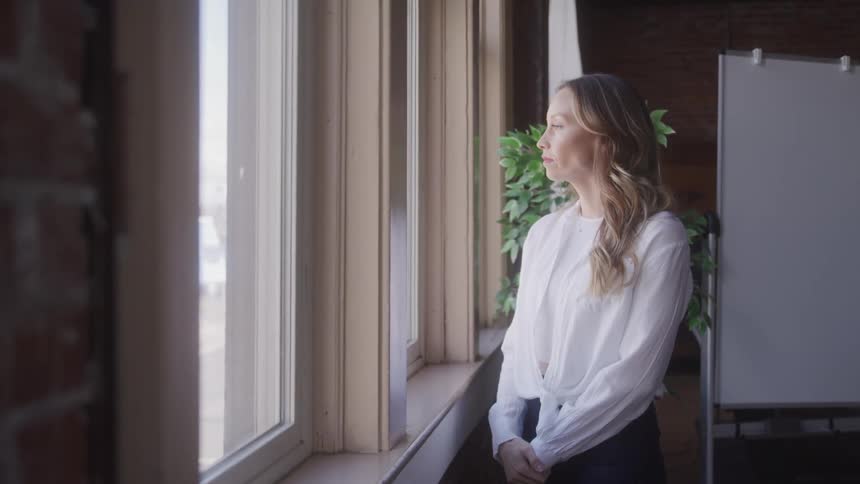 A woman in a white blouse stands in a brightly lit room looking out of a window on a sunny day