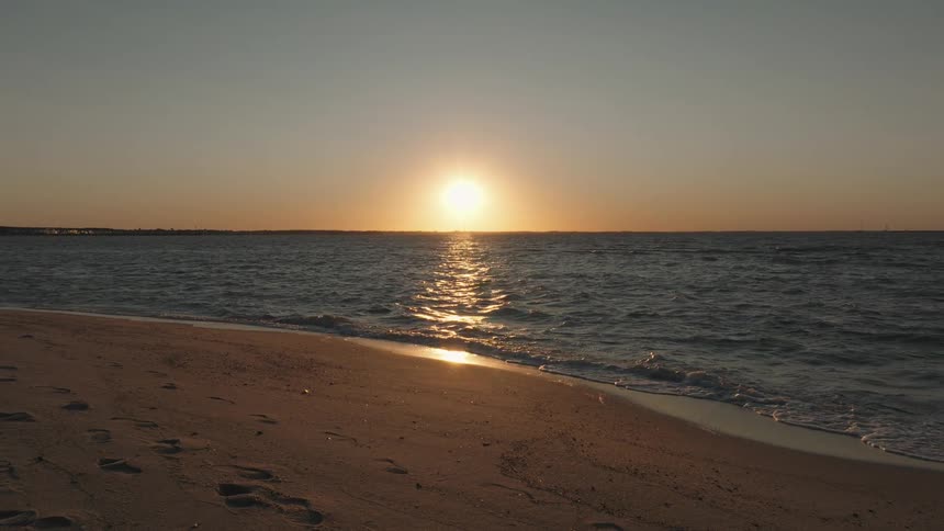 The sun sets over a calm ocean with gentle waves lapping against a sandy beach creating a tranquil and peaceful scene