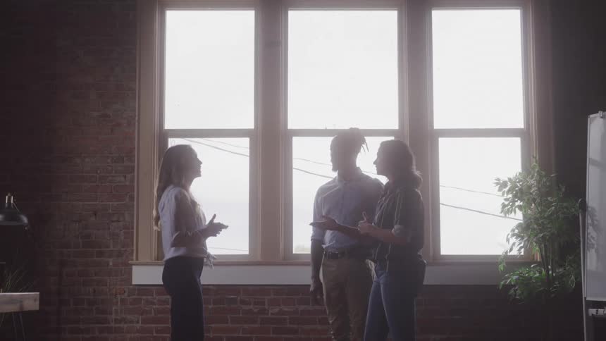 Three people are standing and talking near a large window in what appears to be an office or studio space