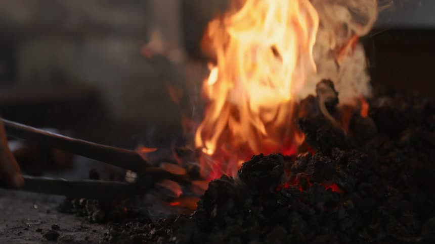 A closeup shows a blacksmith tending to flaming coals in a forge