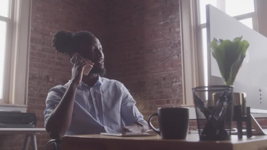 A man with braided hair in a bun talks on a smartphone while sitting at a table near a window in a room with a brick wall