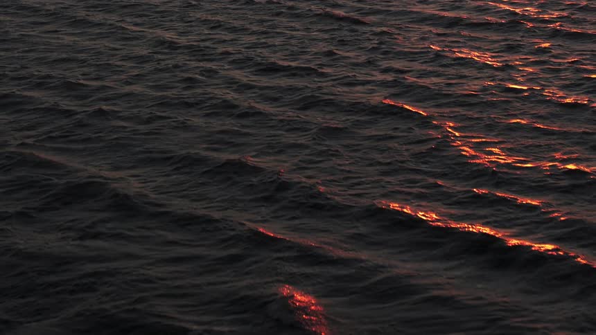 An aerial view shows the ocean surface at sunset with small waves reflecting the reddish sunlight on the dark water below