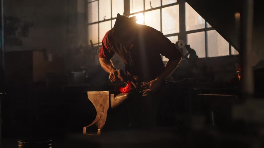 A man is hammering red hot metal on his anvil near the forge