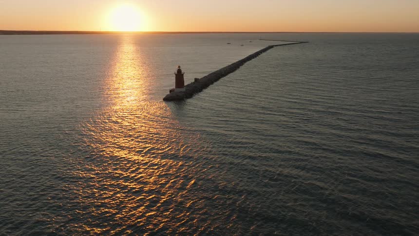 An aerial view of a lighthouse standing at the end of a long rocky breakwater extending into the ocean with the suns reflection shimmering on the water