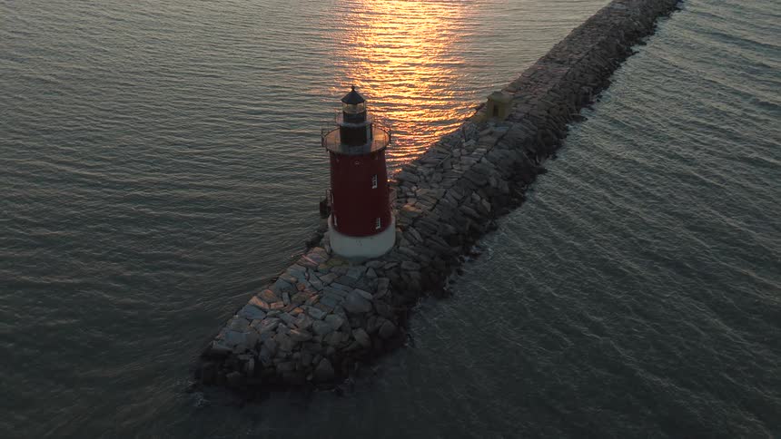 An aerial view of a red and white lighthouse standing at the end of a long rocky breakwater extending into the dark ocean