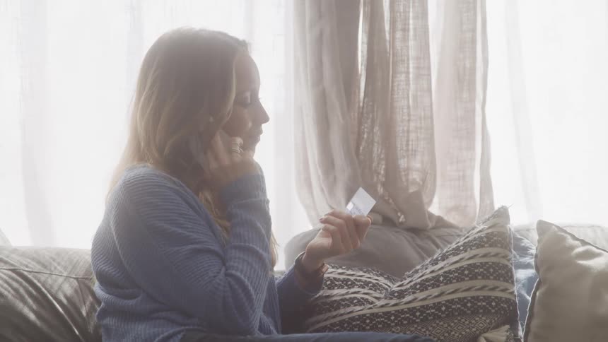 Woman sitting on her couch looks at her credit card while talking on the phone