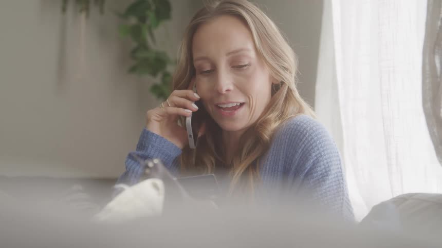 A woman with long blonde hair talks on a cell phone while seated indoors