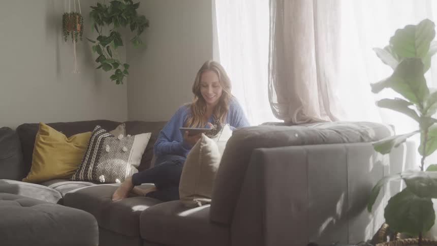 A woman is smiling while sitting on the couch and browsing the internet on a tablet