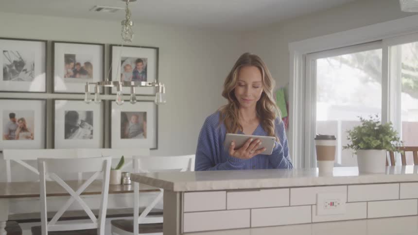 A woman with long blonde hair uses a tablet in a well lit kitchen with white cabinets and framed photos on the wall