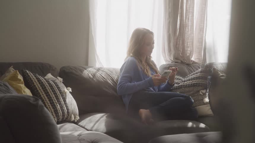 Blonde woman sitting in the living room enters credit card information onto a smartphone