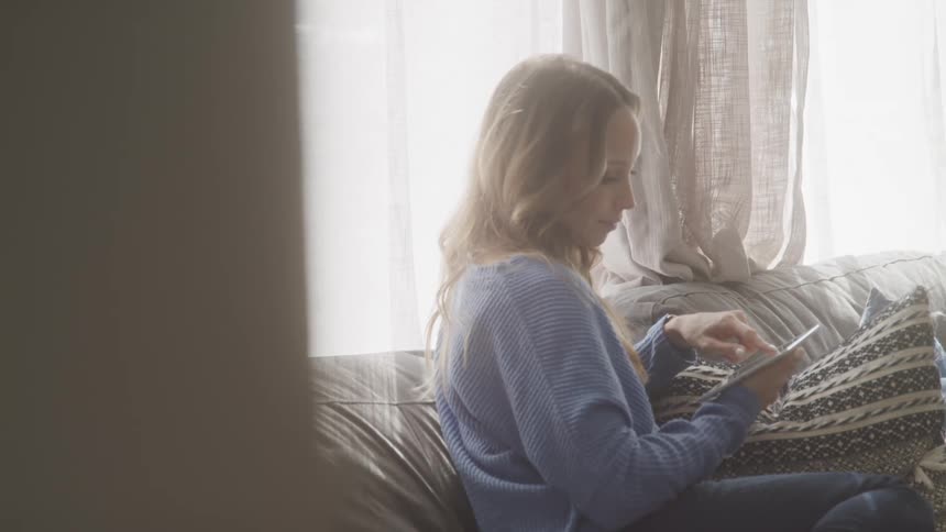 A woman with blonde hair and a blue sweater sits on a couch by the window looking at a tablet screen
