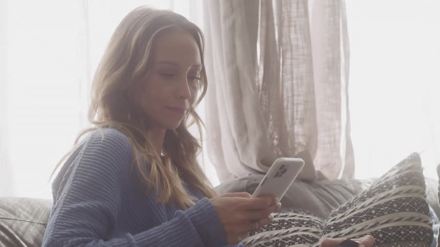 A woman in a blue sweater sits on a couch near a window using her phone in bright sunlight