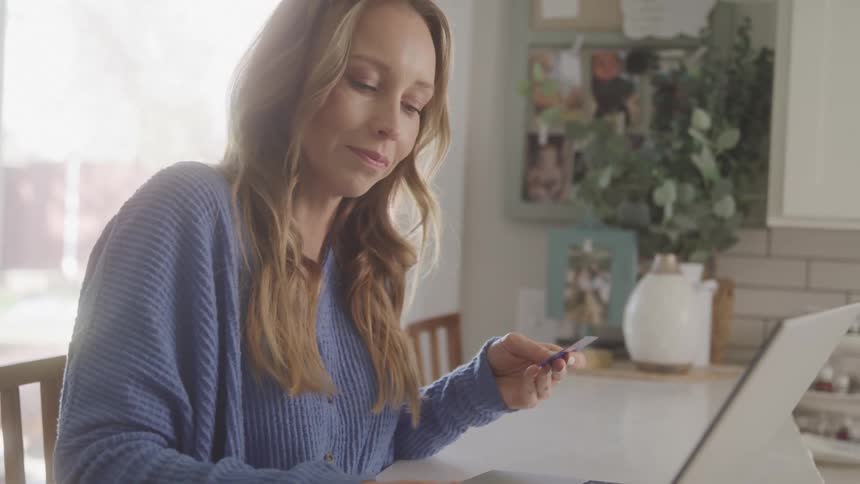 A blonde woman sits indoors and enters credit card information into a laptop computer