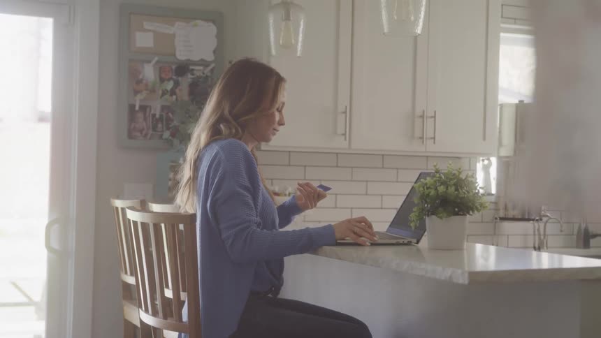 A woman sits in her kitchen on a sunny day holding a credit card in one hand while using a laptop