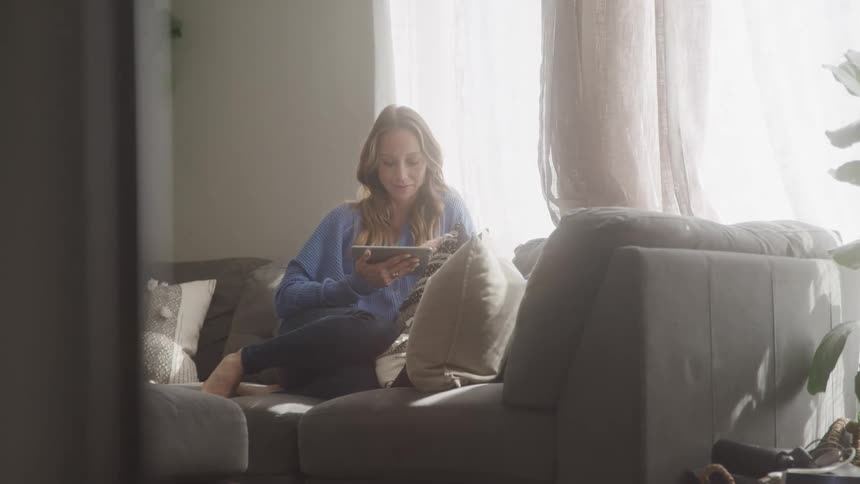 A woman with long blonde hair sits on a couch looking at a tablet in a brightly lit room with a potted plant and natural light coming through a window