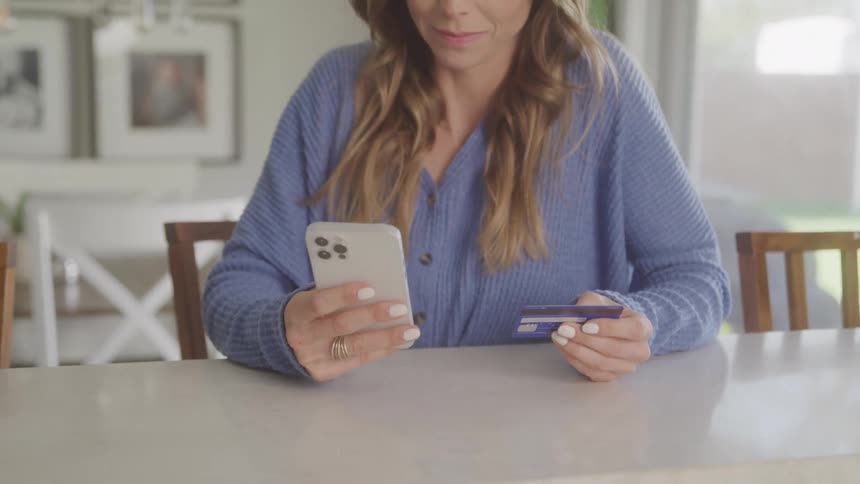 A woman with long wavy blonde hair is sitting at a table looking at her phone and holding a credit card likely making an online purchase