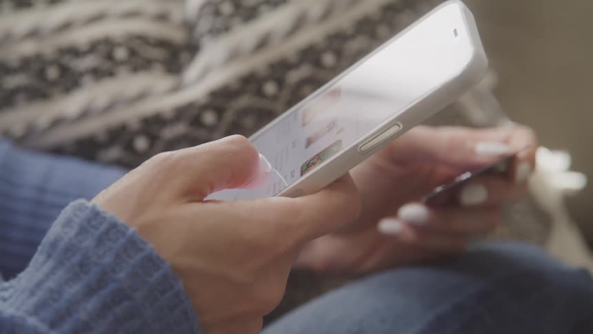 A closeup shows a woman browsing social media on her phone while sitting on a couch at home during the day