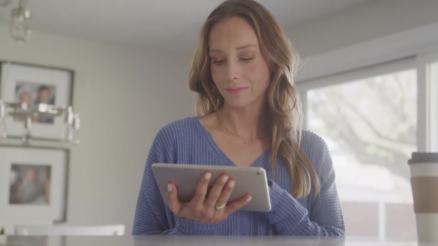 A woman with long blonde hair uses a tablet in a well lit room with picture frames on the wall