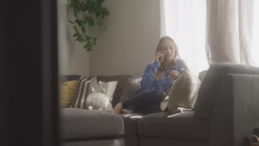 A woman with long blonde hair sits on a couch talking on a cell phone in a brightly lit room with a potted plant and natural light coming through a window