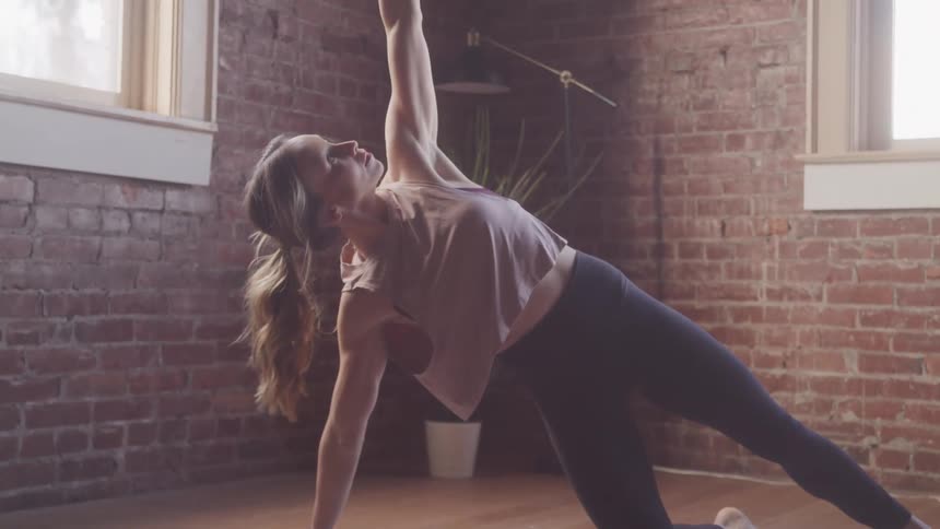 A woman stretches on a mat in a sunlit room with a brick wall and large windows on a bright day