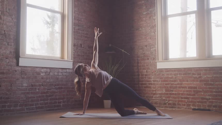 Woman balances in a yoga pose on a yoga mat placed in a corner between windows