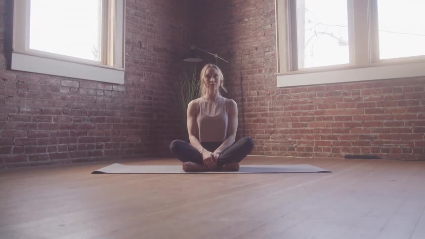 A woman with blonde hair sits cross legged on a yoga mat in a room with exposed brick walls practicing yoga