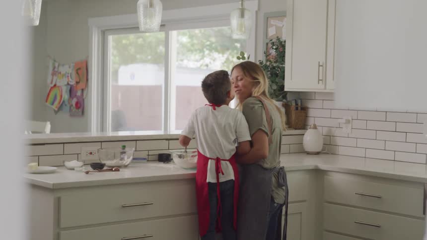 Mother and son both wearing aprons are smiling and kissing each other as they prepare something in the kitchen