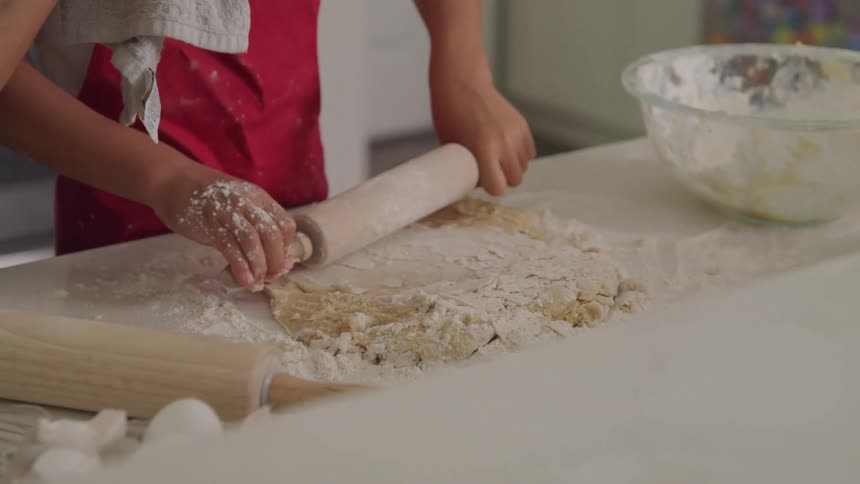 A boy in a red apron uses a rolling pin to flatten dough on a countertop