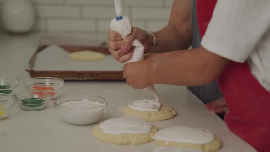 A woman helps a boy in a red apron apply frosted topping to cookies while cooking at home