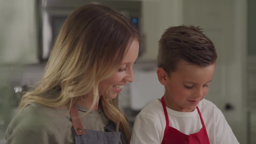 A mother and her young son both wearing aprons work together on a task in a kitchen setting with the mother smiling