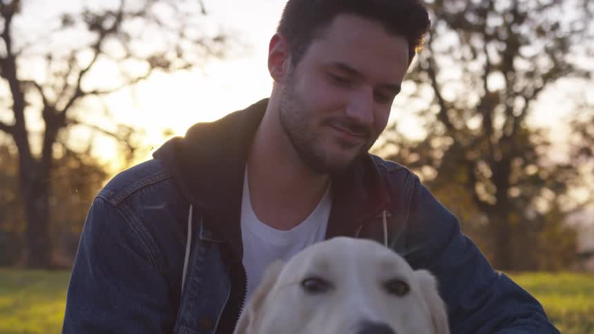 A man with light colored hair pets a dog in a sunny outdoor setting with trees in the background