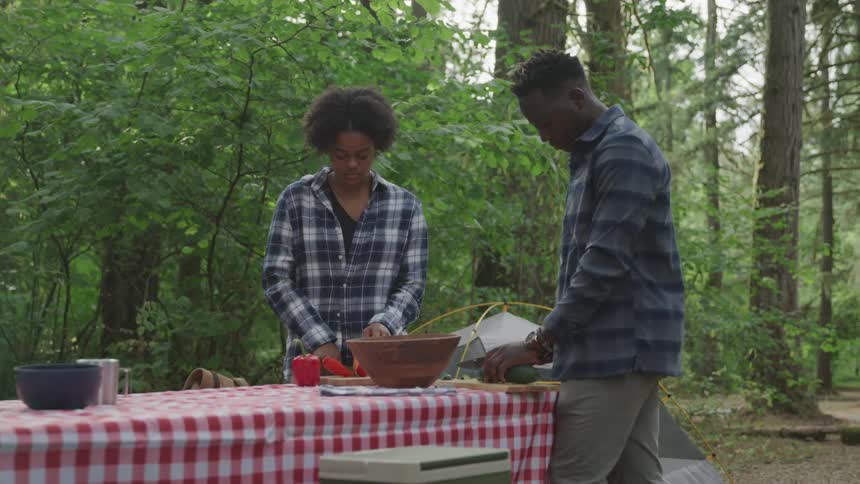 2 boys cutting food at a table in the middle of the woods