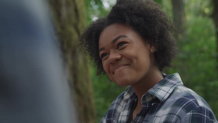 The face of a young woman who is smiling subtly looking at someone who is in front of her and there are trees behind her